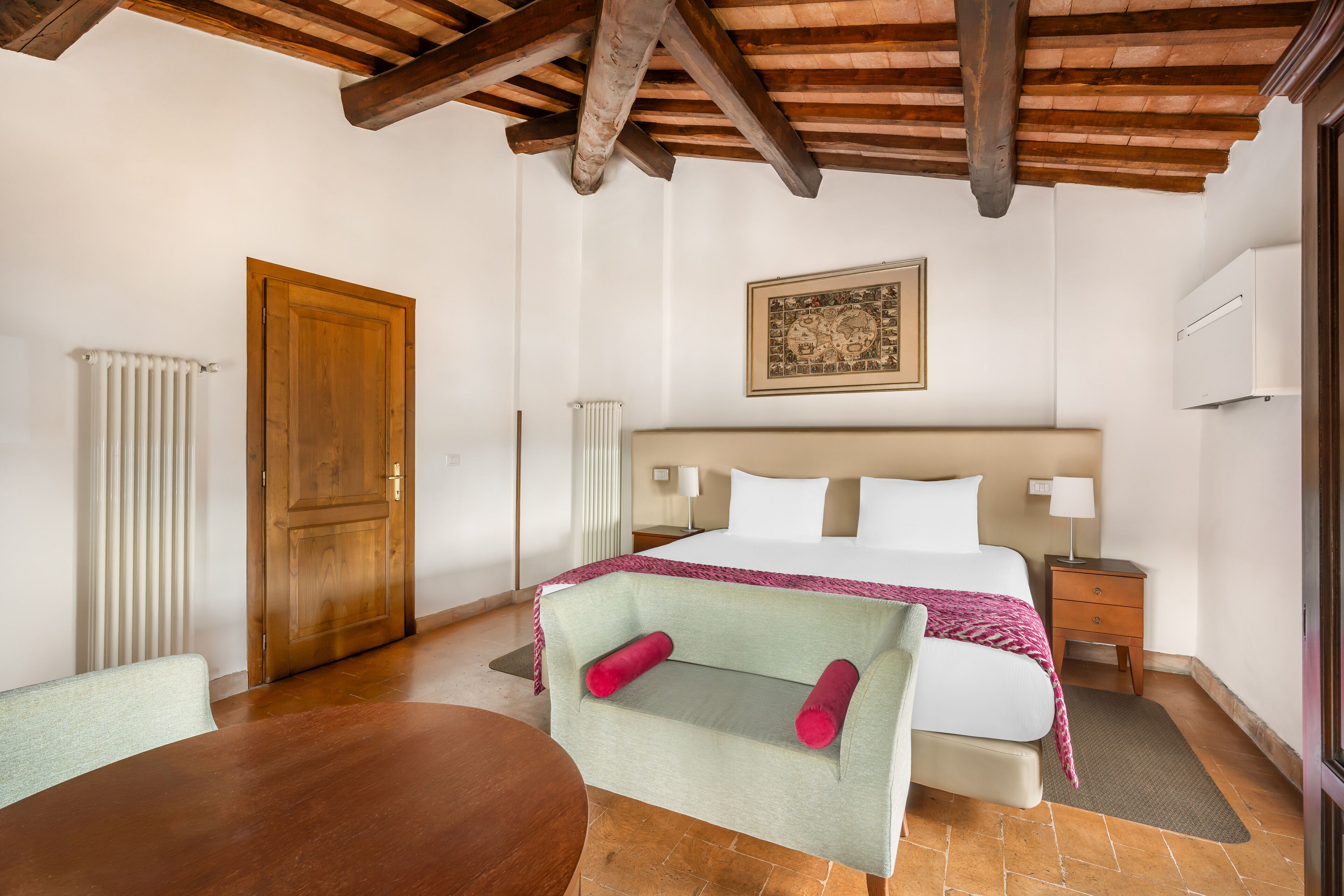 A hotel room with a bed, a wooden ceiling, a radiator, a door, a nightstand, a table, and a chair.