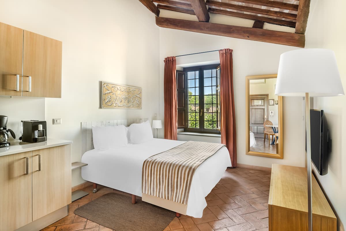 Un dormitorio con cama, escritorio, televisión, ventana con cortinas y una zona de cocina con armarios y cafetera.