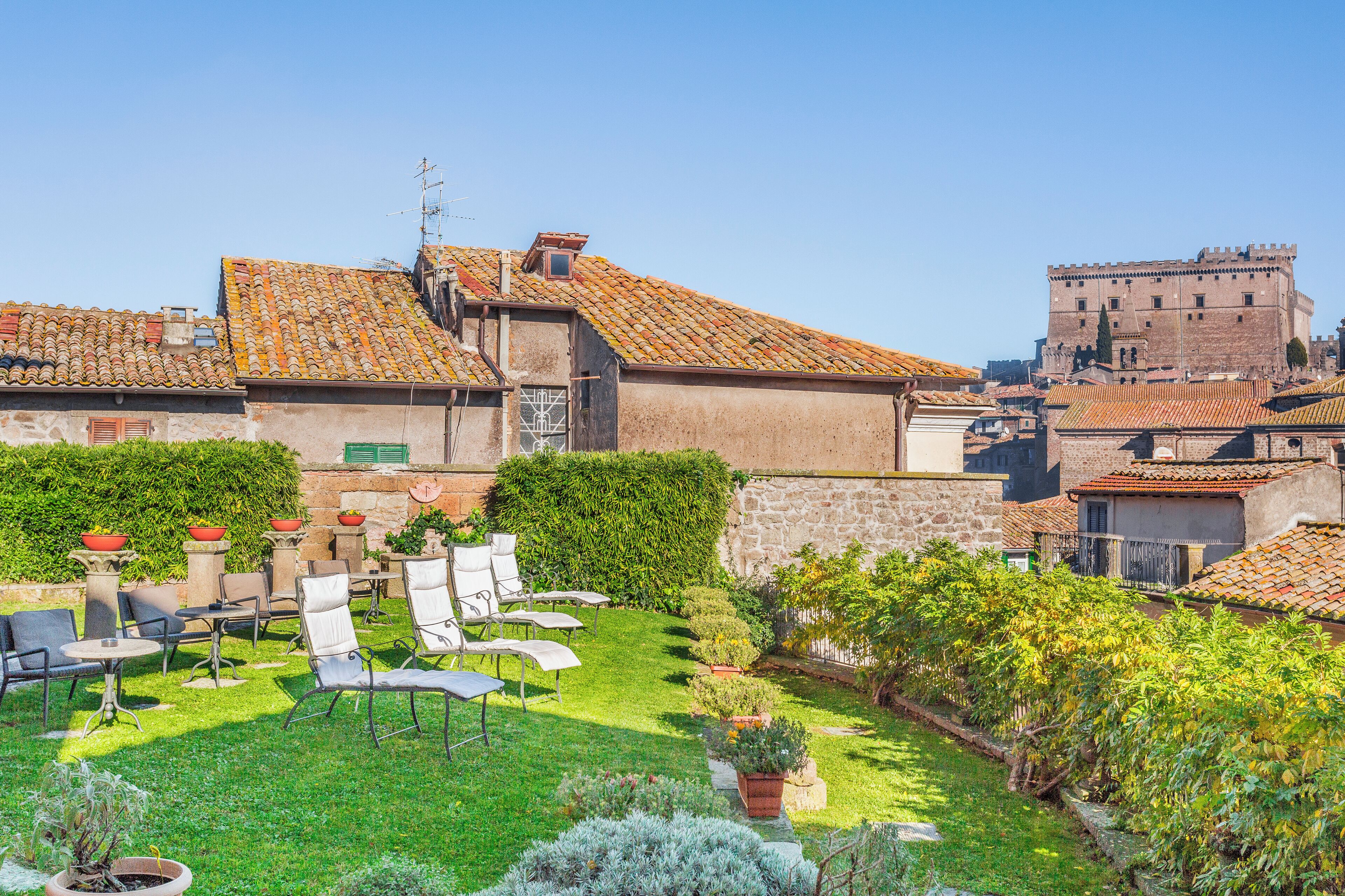 A courtyard with outdoor seating, a stone wall, and a large building with a tiled roof in the background.