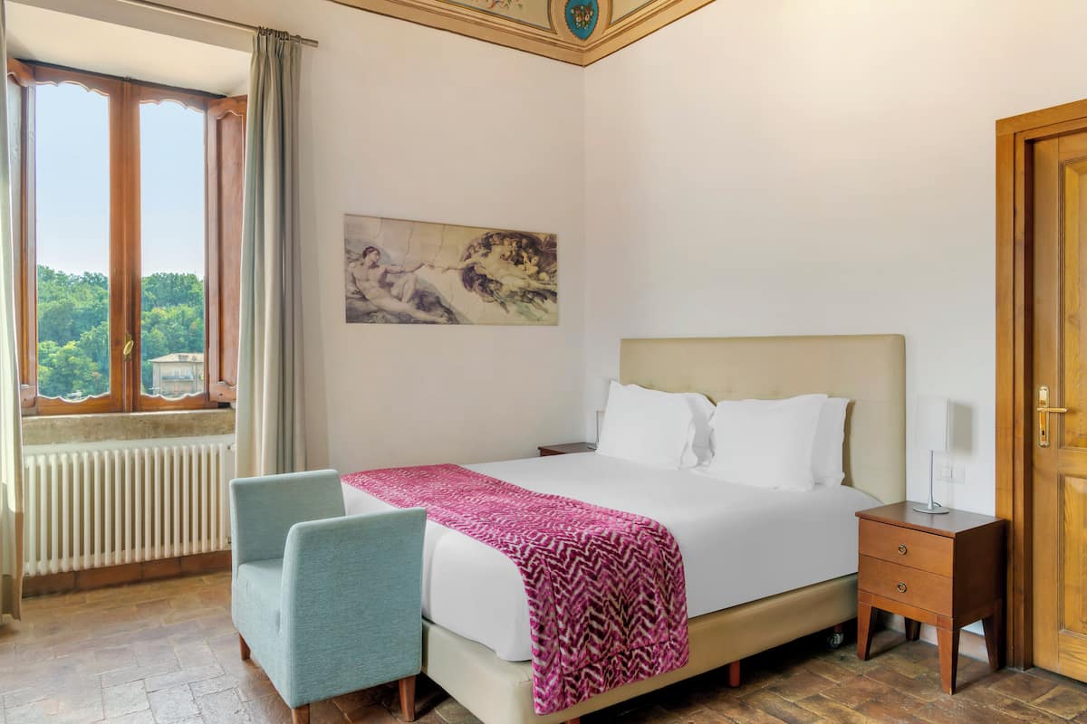 Una habitación de hotel con una cama, una silla, una mesita de noche, una ventana con cortinas y un cuadro en la pared.