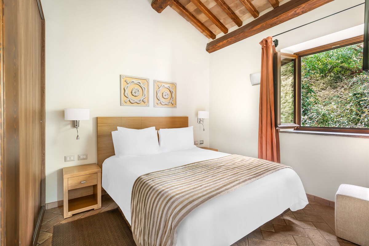 A hotel room with a large bed, wooden headboard, two wall-mounted lamps, a window with curtains, and a small bedside table.