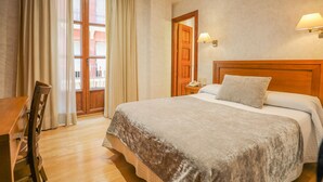 A hotel room with a bed, wooden headboard, bedside table, lamp, and a view of the outside through a window with curtains. - Hotel Granada Centro (Granada)