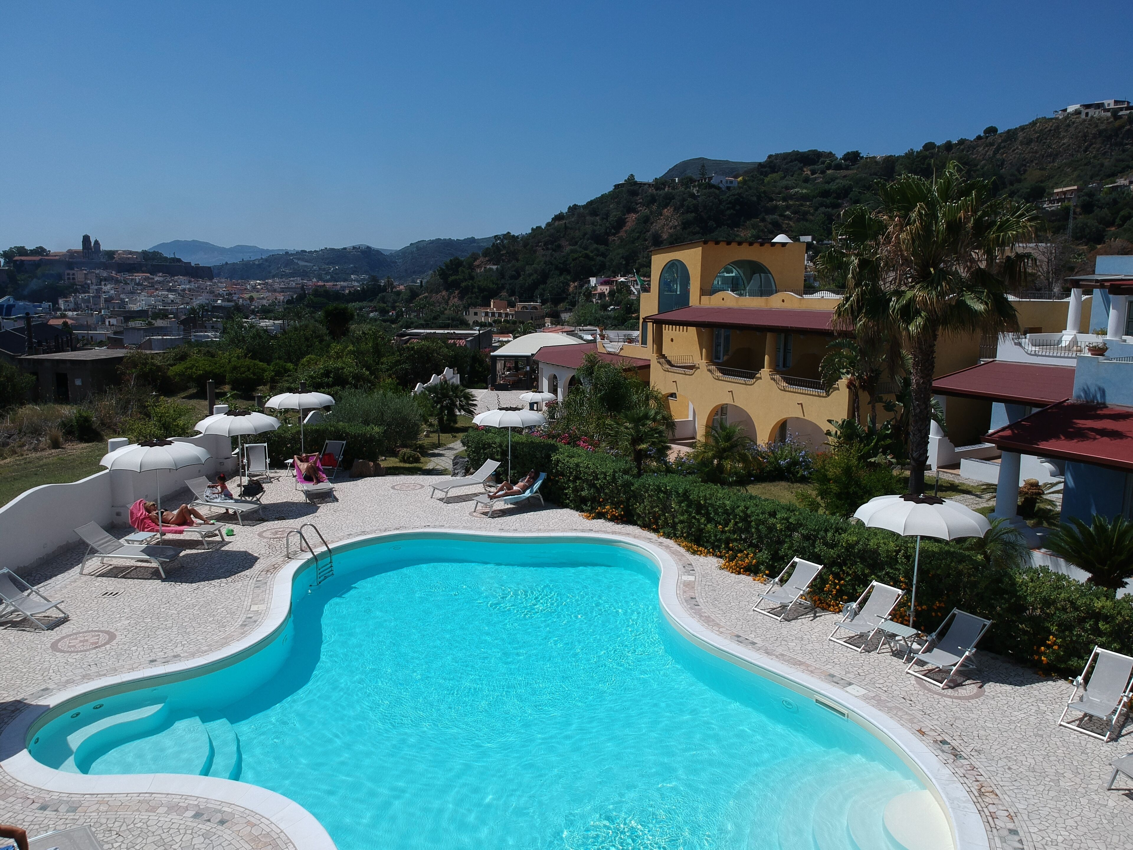 Une piscine entourée de verdure et de bâtiments, avec des chaises longues et des parasols.