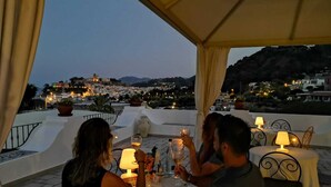 Una terraza con una mesa preparada para tres personas, con vistas a un paisaje urbano al anochecer.