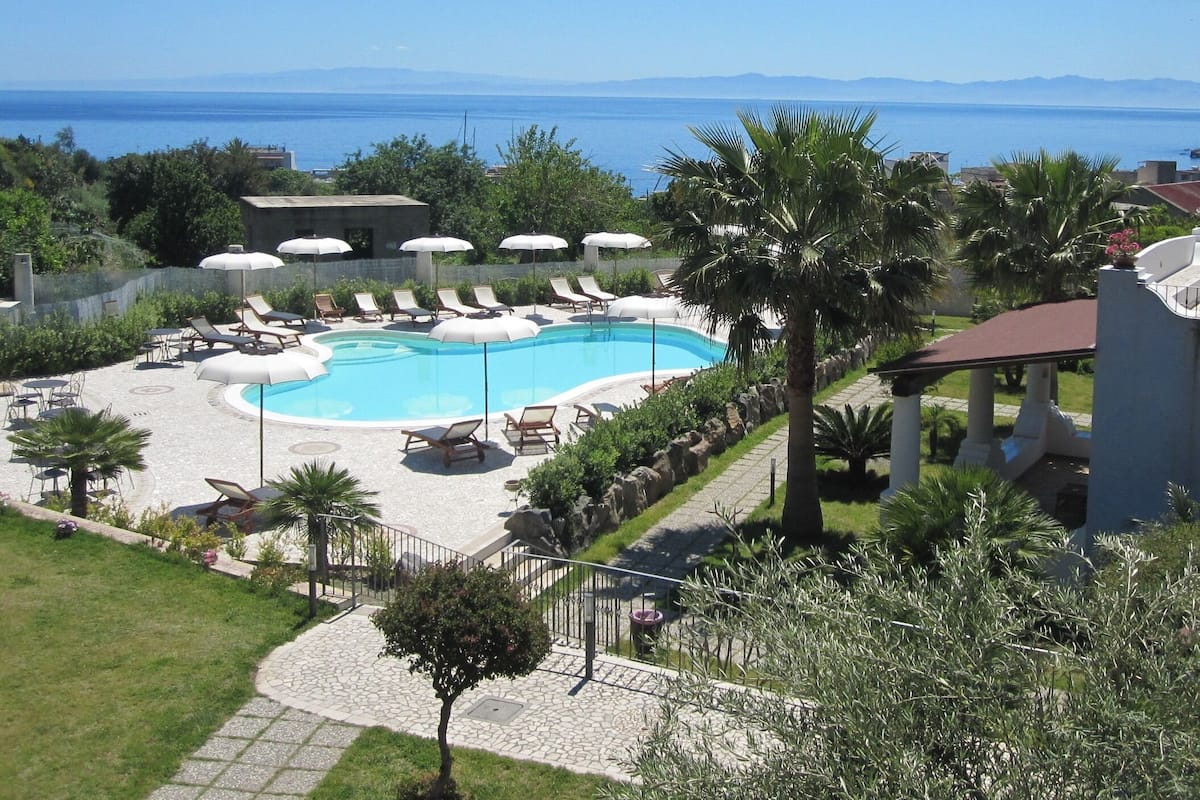 A pool area with lounge chairs and umbrellas, surrounded by greenery and a view of the sea.