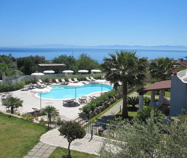 A pool area with lounge chairs and umbrellas, surrounded by greenery and a view of the sea.