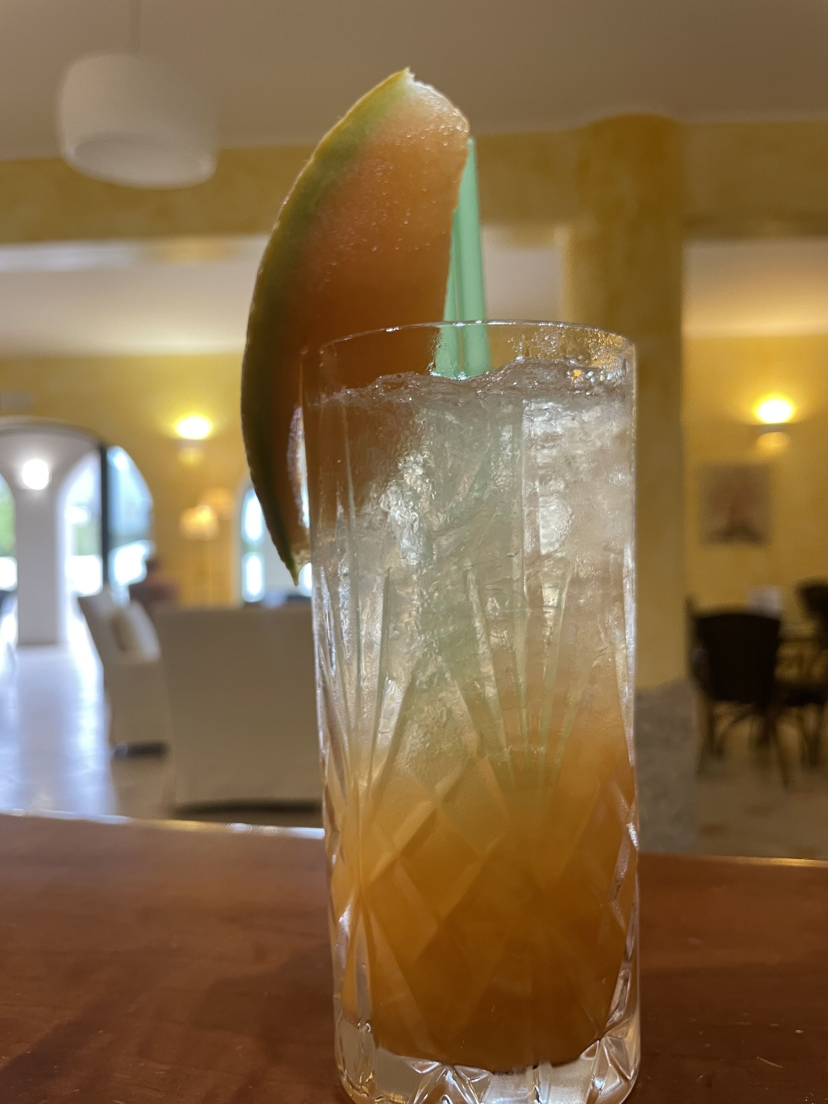 A tall glass with a lime wedge and a straw, filled with a yellowish beverage, on a wooden table in a room with a yellow wall and a window in the background.