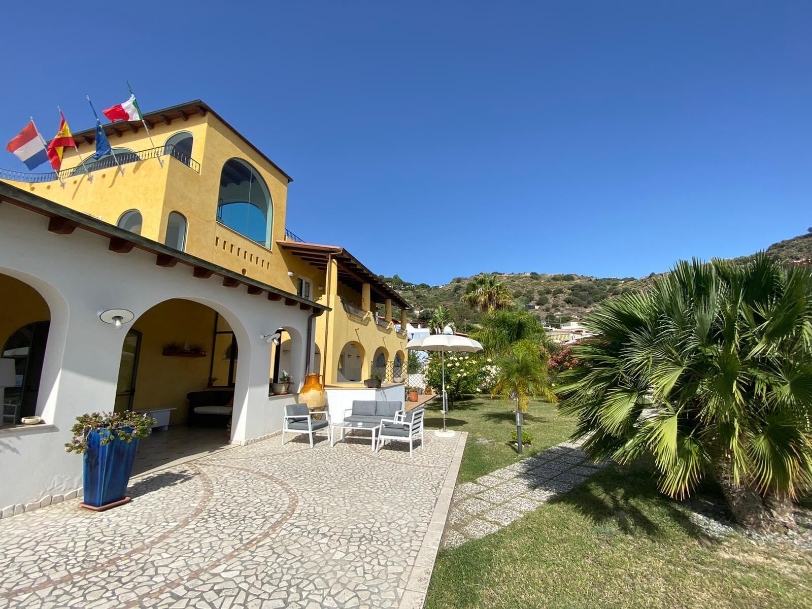 Un bâtiment jaune orné d’arches et de drapeaux, une terrasse avec des chaises et une table, et un palmier.