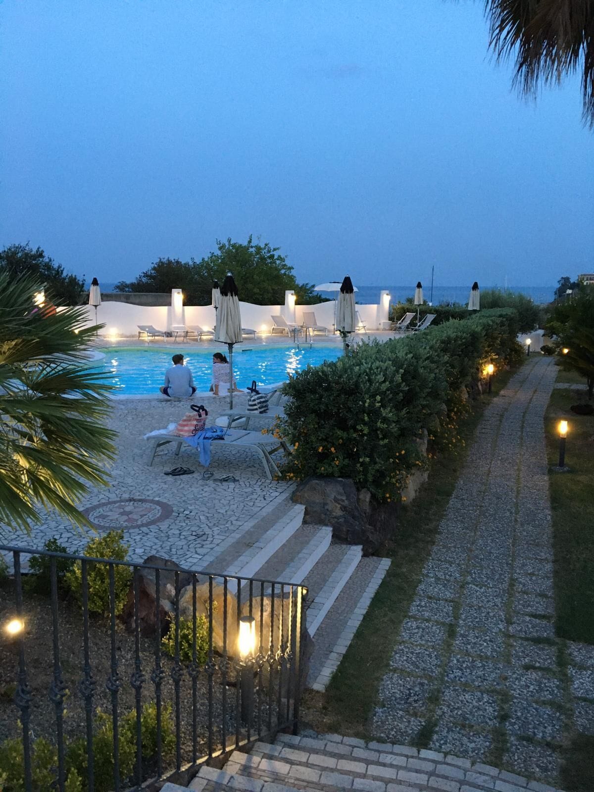 A poolside area with lounge chairs and umbrellas, a paved walkway, and a stone wall.