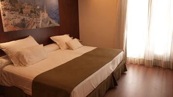 A neatly made bed with a brown bedspread, white linens, and two pillows. A large painting of a coastal town is on the wall above the bed.