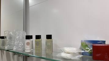 Un baño con un estante de vidrio que sostiene varios artículos de tocador, como botellas de champú y loción corporal, una cajita de pañuelos y una caja roja.