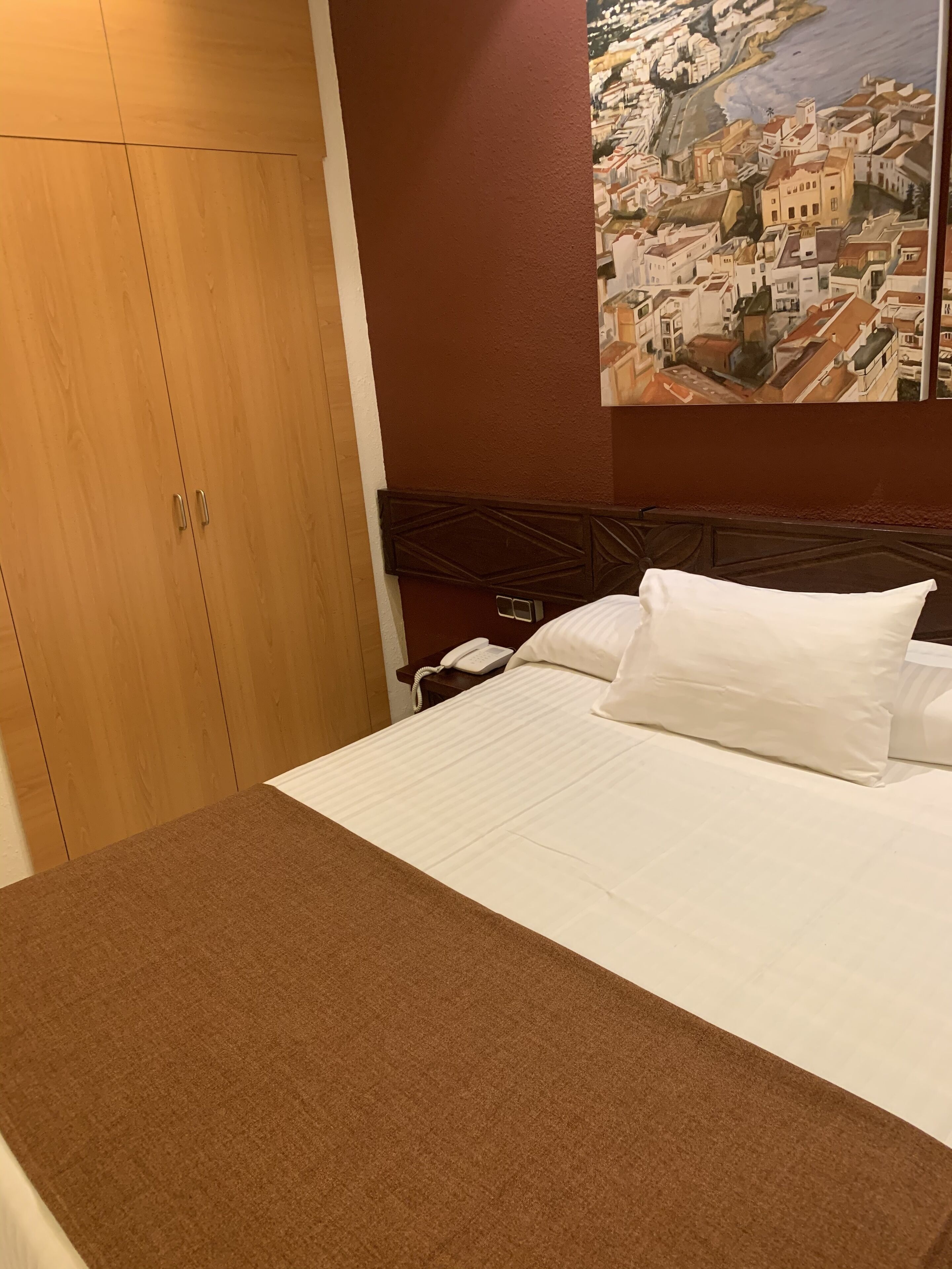 A hotel room with a bed, a wooden wardrobe, and a painting of a coastal town.