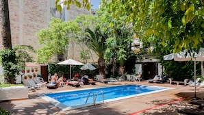 Zona de piscina con sillas de descanso y sombrillas, rodeada de árboles y un edificio al fondo.