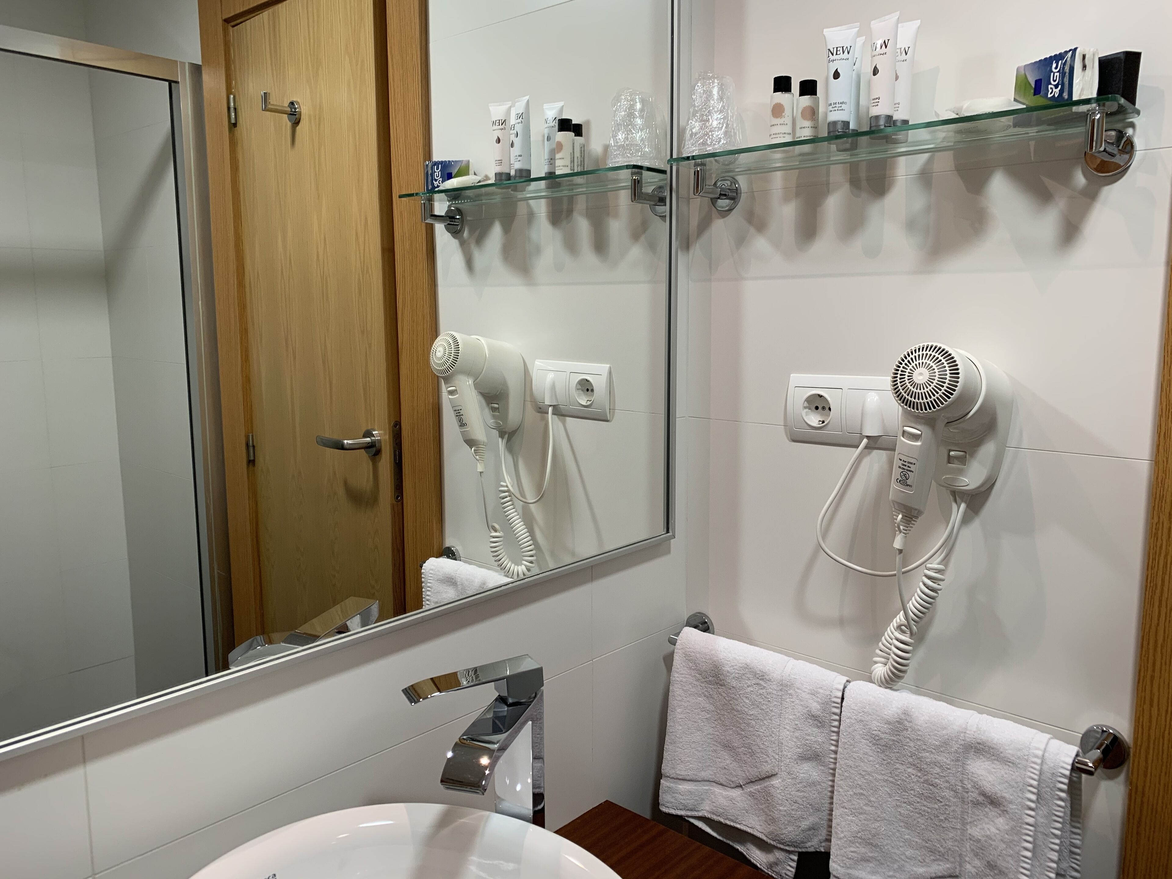 A bathroom with a white sink, a mirror, a glass shelf with toiletries, and two hair dryers plugged in.