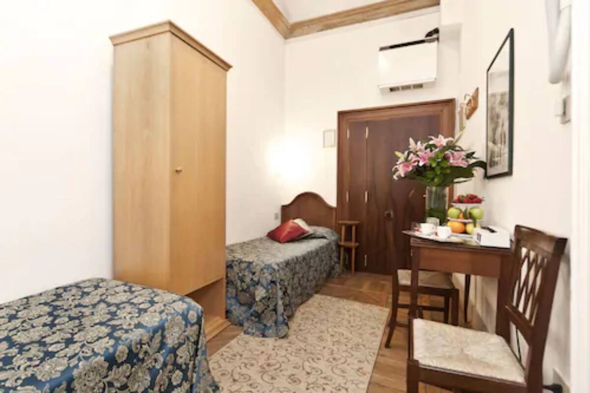 A bedroom with two beds, a wooden wardrobe, a desk with a chair, and a small table with a vase of flowers.