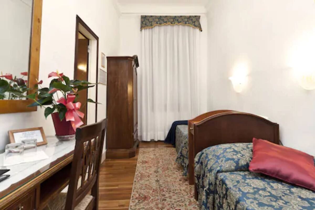 A hotel room with a wooden desk, a chair, a bed with floral bedding, a nightstand, a mirror, and a window with curtains.
