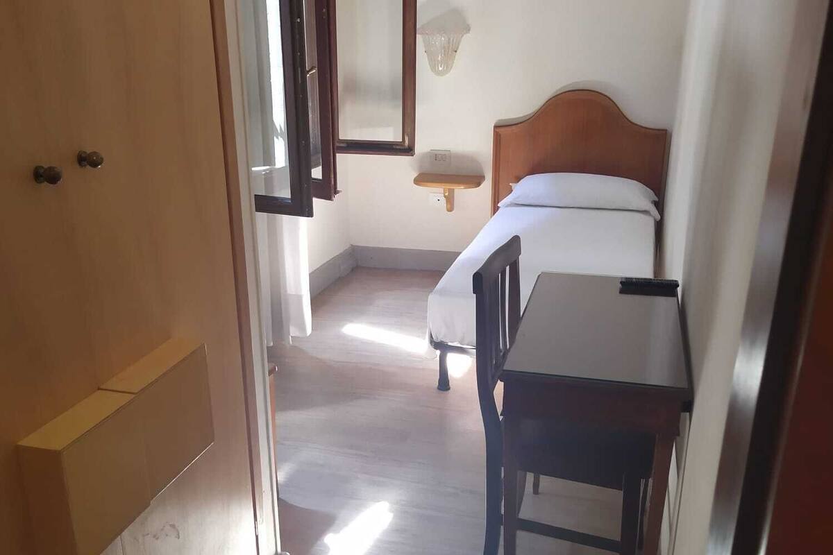 A bedroom with a bed, bedside table, and a chair. There is a window with a view of the outside.