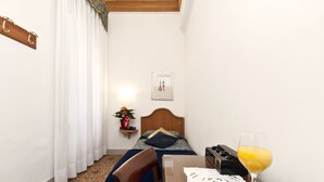 A hotel room with a bed, a bedside table with a glass of orange juice, a chair, a small table with pastries, and a wall-mounted shelf with a plant. - Hotel Alla Fava (Venice)