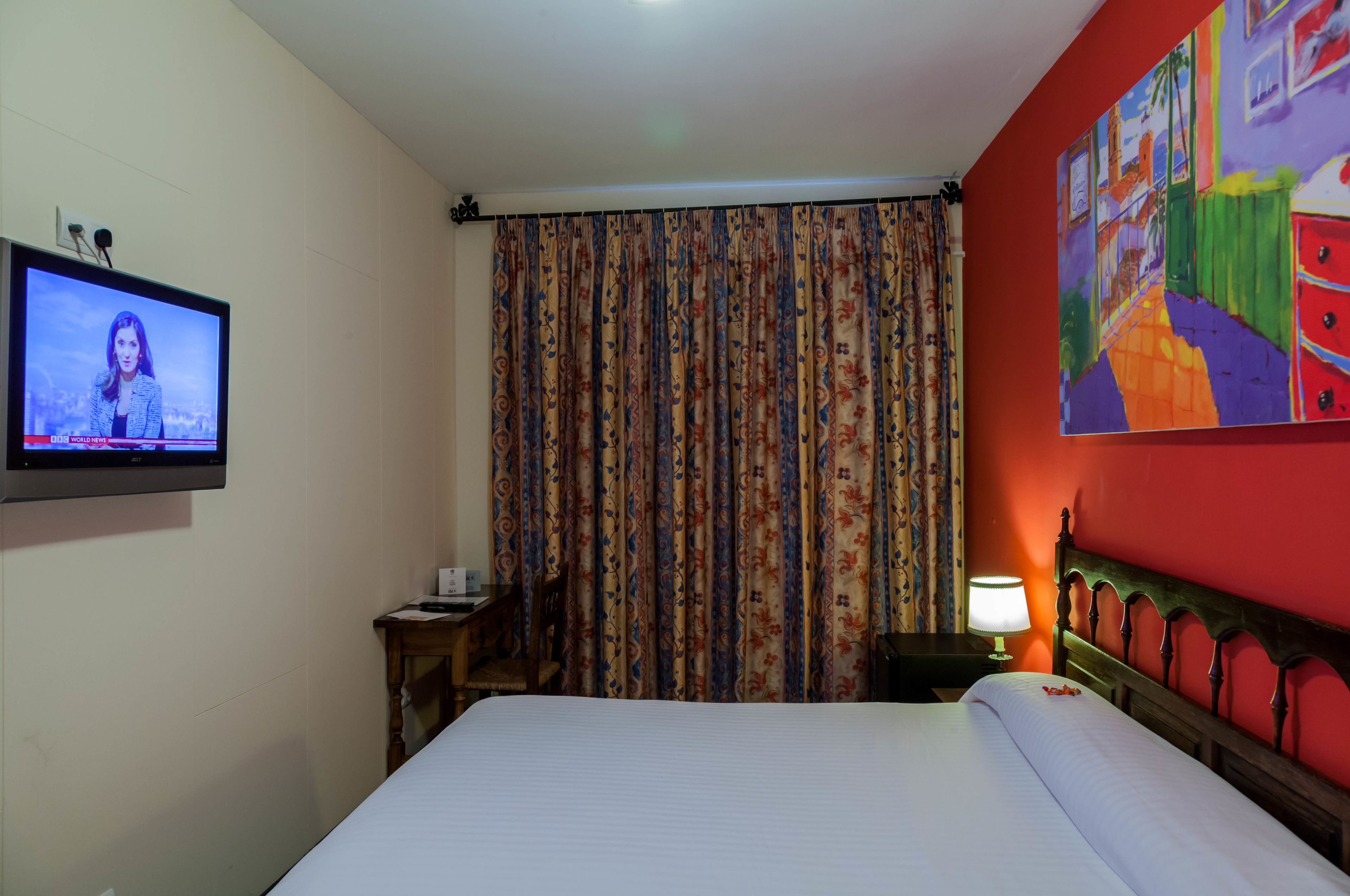 Una habitación de hotel con una cama, un televisor en la pared, un escritorio con una lámpara y un cuadro de colores en la pared.