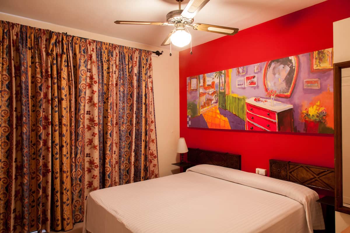 A hotel room with a bed, curtains, a ceiling fan, a painting on the wall, and a nightstand.