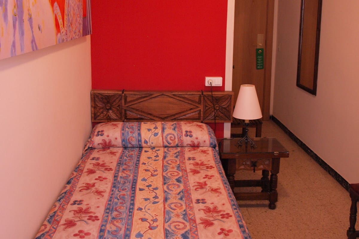 A hotel room with a red wall, a bed with a floral bedspread, a wooden headboard, a bedside table with a lamp, and a painting on the wall.