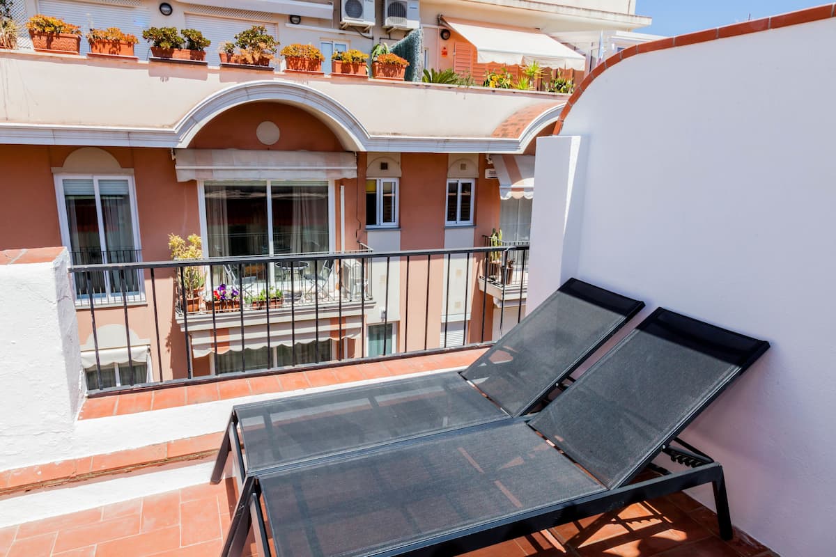 A balcony with two sun loungers, a railing, and a view of a building with potted plants and a door.