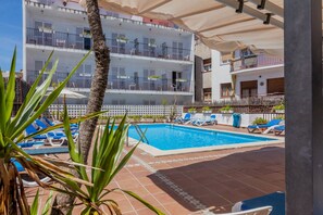 A pool area with blue lounge chairs, a palm tree, and a multi-story building with balconies. - Hotel El Cid (Sitges)