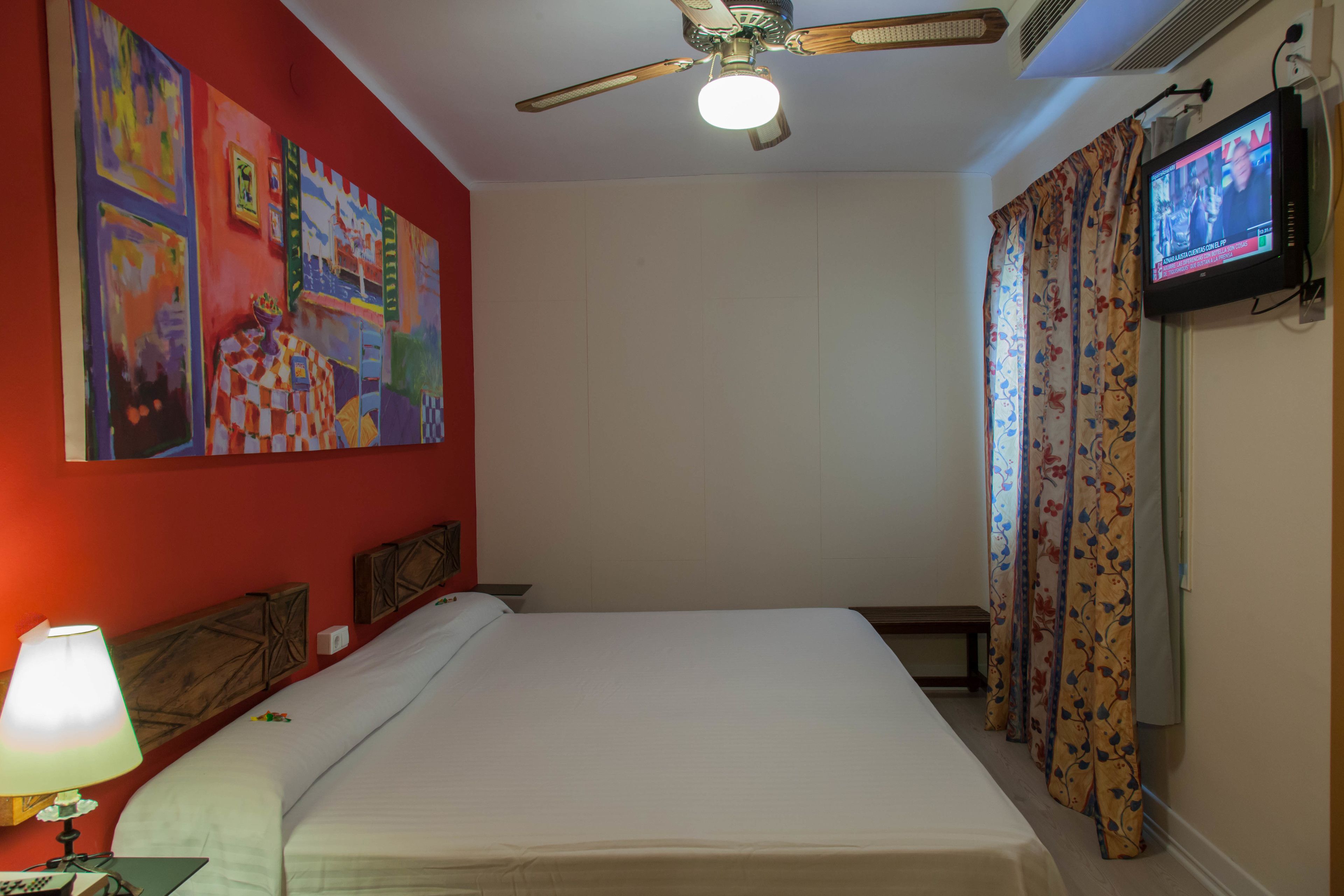 Una habitación de hotel con una cama, un ventilador de techo, un televisor y una pared decorada con obras de arte coloridas.