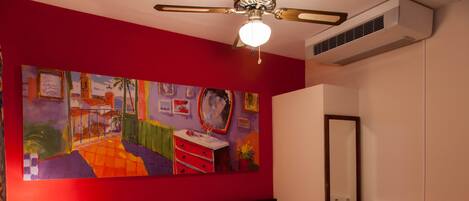 A hotel room with a large painting on the wall, a bed, a nightstand, a lamp, a mirror, and a shelf with a clock.