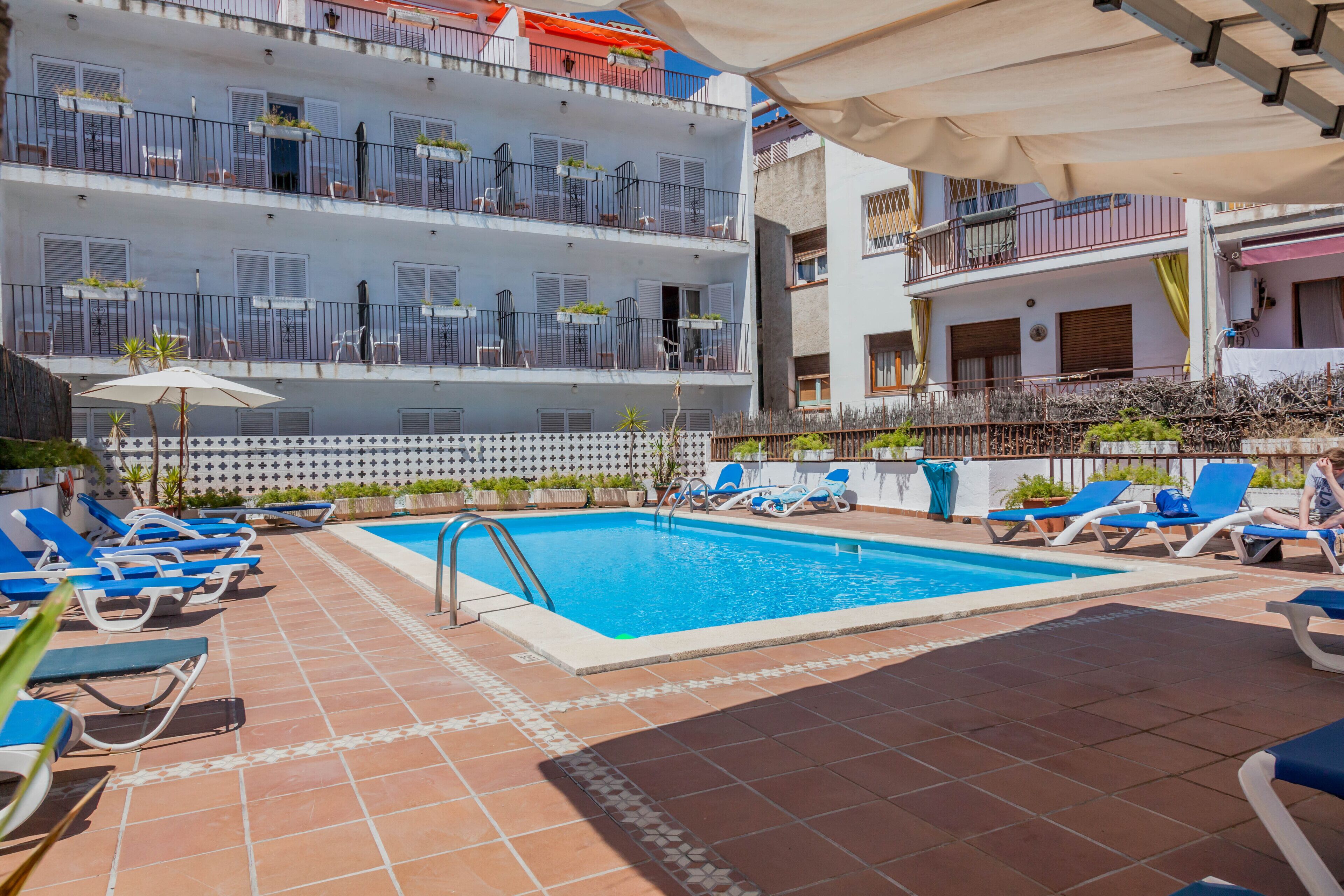 Un área de piscina en la azotea con sillas reclinables azules, una piscina y un edificio con balcones.
