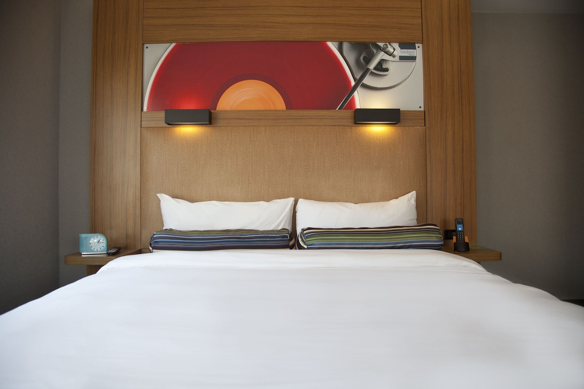 A neatly made bed with white linens, two pillows, and a striped blanket. There is a small bedside table with a blue alarm clock and a remote control. A wall-mounted light fixture is above the bed.
