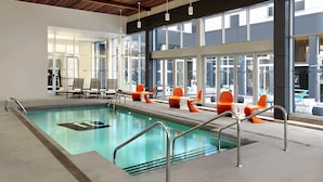 An indoor swimming pool with clear water, surrounded by orange chairs and tables, large windows, and a wooden ceiling. - Aloft by Marriott Montreal Airport (Dorval)