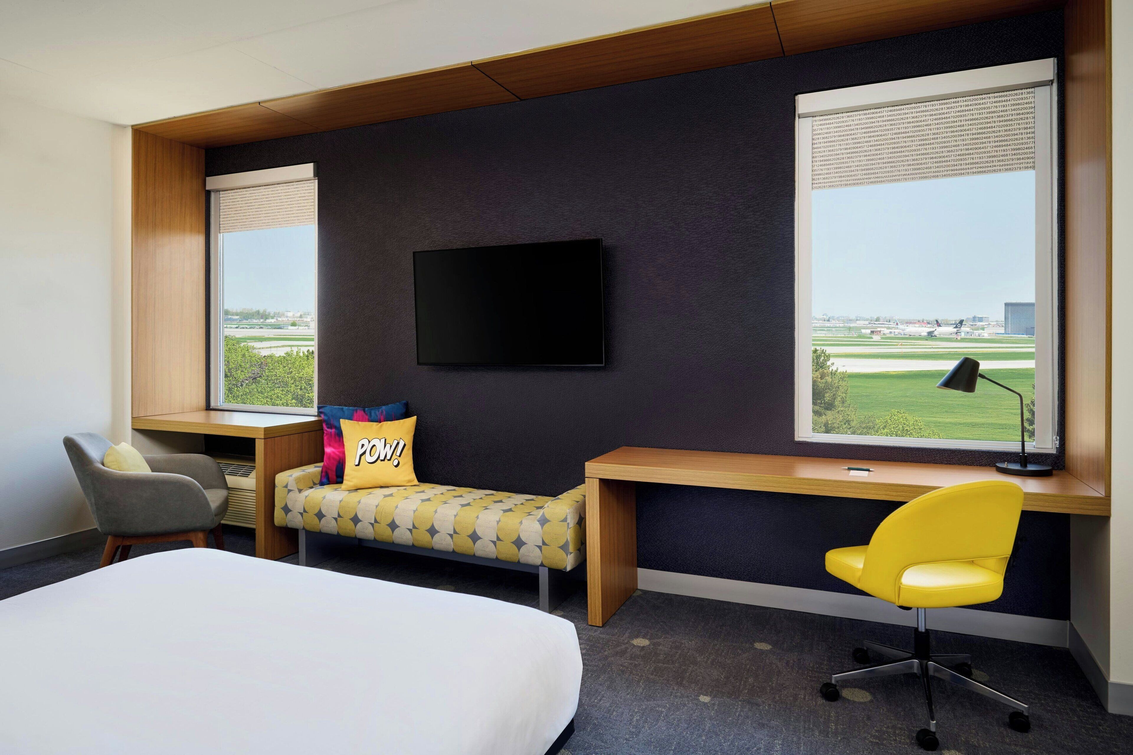 A hotel room with a bed, a desk with a chair, a TV mounted on the wall, and large windows offering a view of the airport.