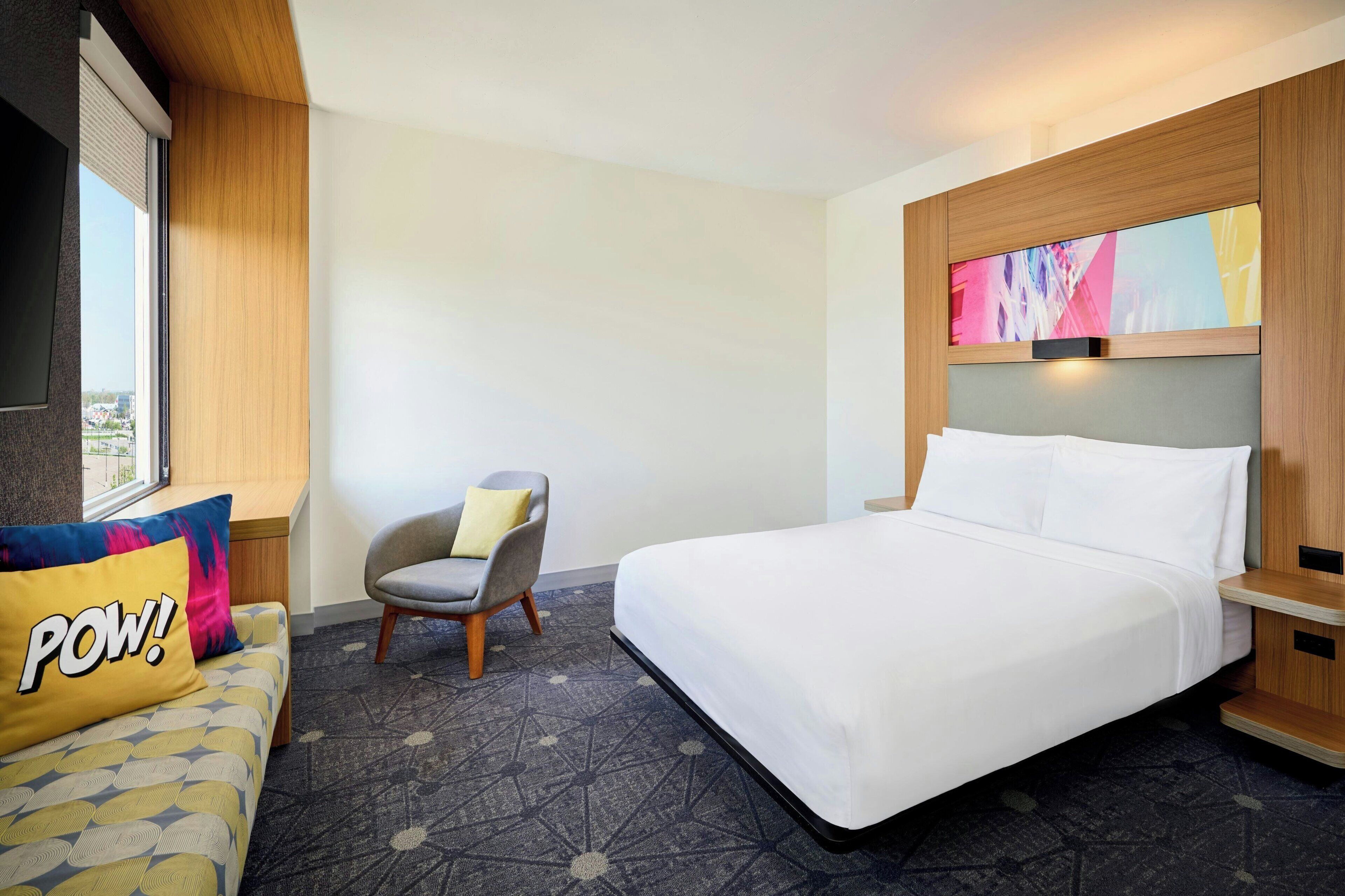 A hotel room with a bed, a chair, a sofa with a 'POW!' pillow, a large window, and a wall-mounted TV.