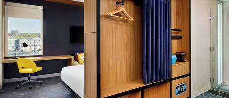 A hotel room with a bed, a desk with a chair, a wardrobe, a shower, and a view of the city.