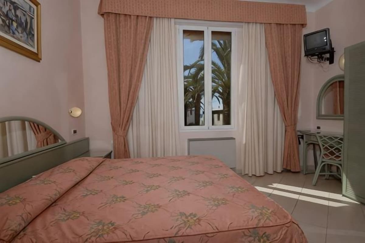 A bedroom with a bed, a window with curtains, a desk with a chair, and a television.