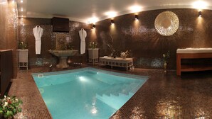 Un espace piscine intérieur luxueux, avec un sol recouvert de mosaïques, un coin salon doté d’un banc et une petite table ornée d’un vase de fleurs.