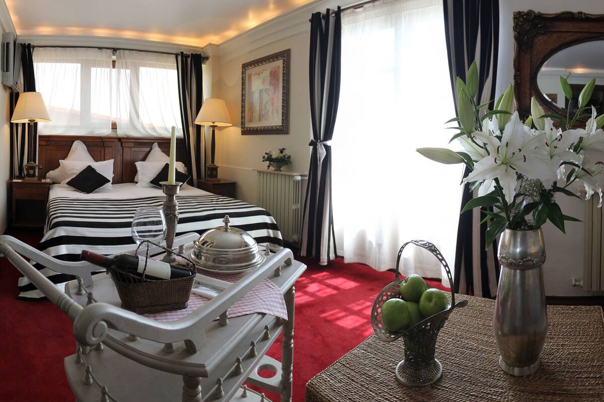 Une chambre d’hôtel comprenant un lit, une table de chevet, une lampe, une chaise et une table ornée d’un vase de fleurs et d’un panier de fruits.