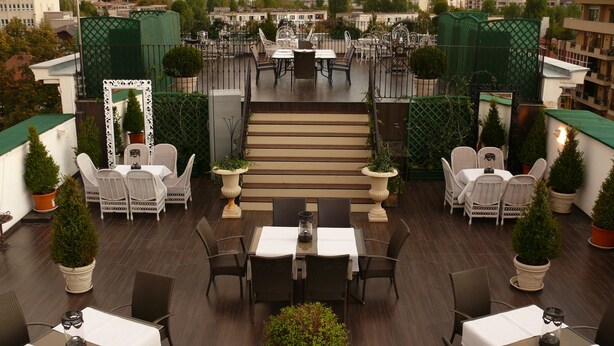 Une terrasse sur le toit, dotée de plusieurs tables et chaises, entourée de plantes en pot et accessible par un escalier menant à un niveau supérieur.