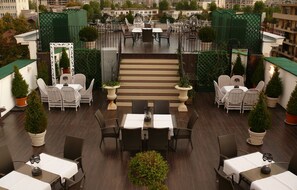 Une terrasse sur le toit, dotée de plusieurs tables et chaises, entourée de plantes en pot et accessible par un escalier menant à un niveau supérieur.