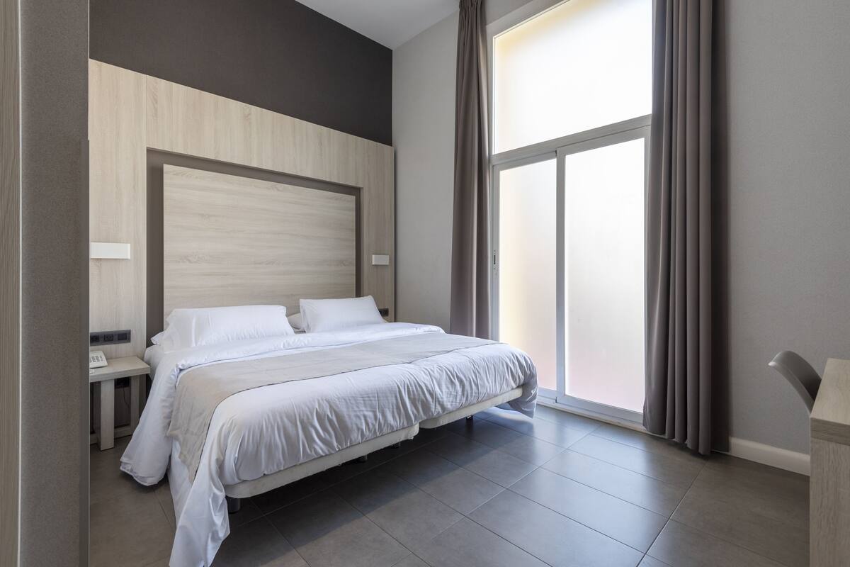 Een moderne hotelkamer met een groot bed, nachtkastje en een groot raam met gordijnen.