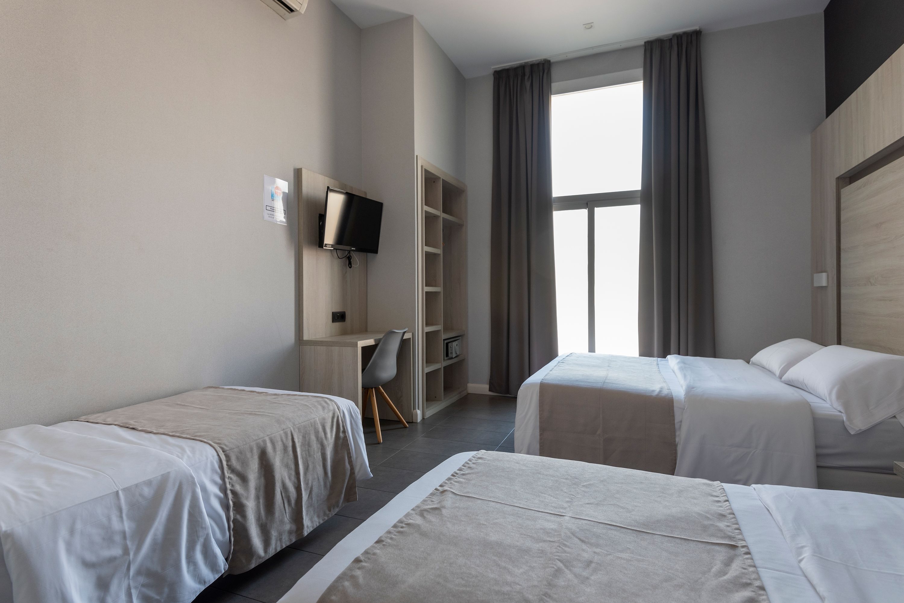 Une chambre d’hôtel avec deux lits simples, un bureau avec une chaise, une télévision et une grande fenêtre avec des rideaux.
