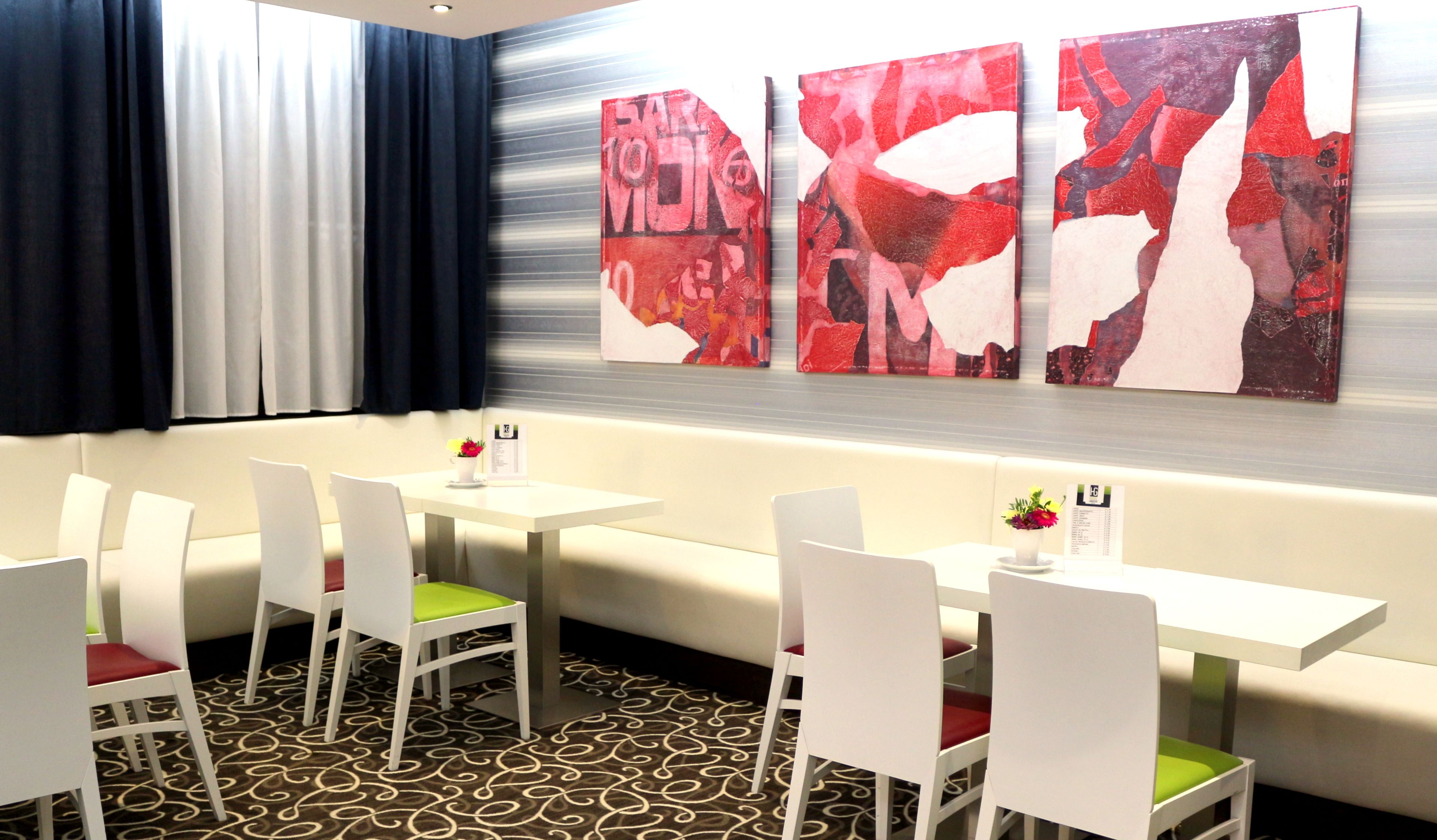 Habitación de hotel con zona de comedor que incluye mesas y sillas blancas, alfombra estampada y arte abstracto en la pared.