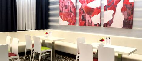 Habitación de hotel con zona de comedor que incluye mesas y sillas blancas, alfombra estampada y arte abstracto en la pared.
