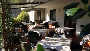 Un espace extérieur aménagé pour les repas, avec des tables et des chaises, entouré de plantes en pot et avec un bâtiment en arrière-plan.