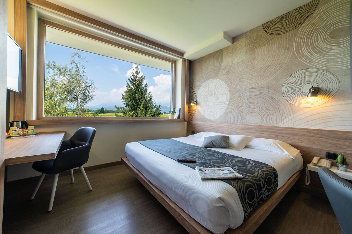 Une chambre d’hôtel moderne dotée d’une grande fenêtre, d’un bureau, d’un lit recouvert d’une couvre-lit à motifs et d’un décor mural original.
