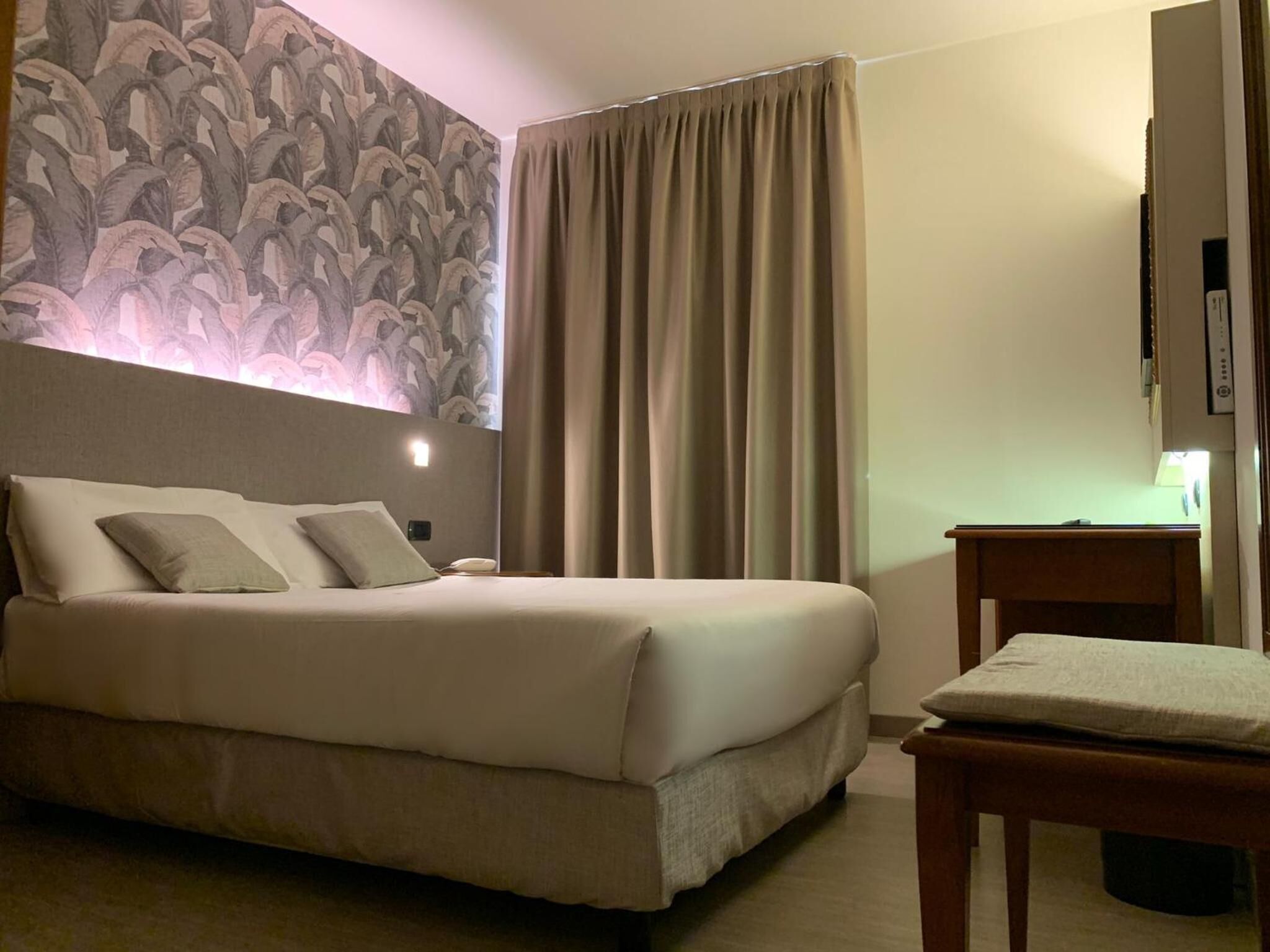 A hotel room with a bed, a desk, a chair, and a wall with a floral pattern.