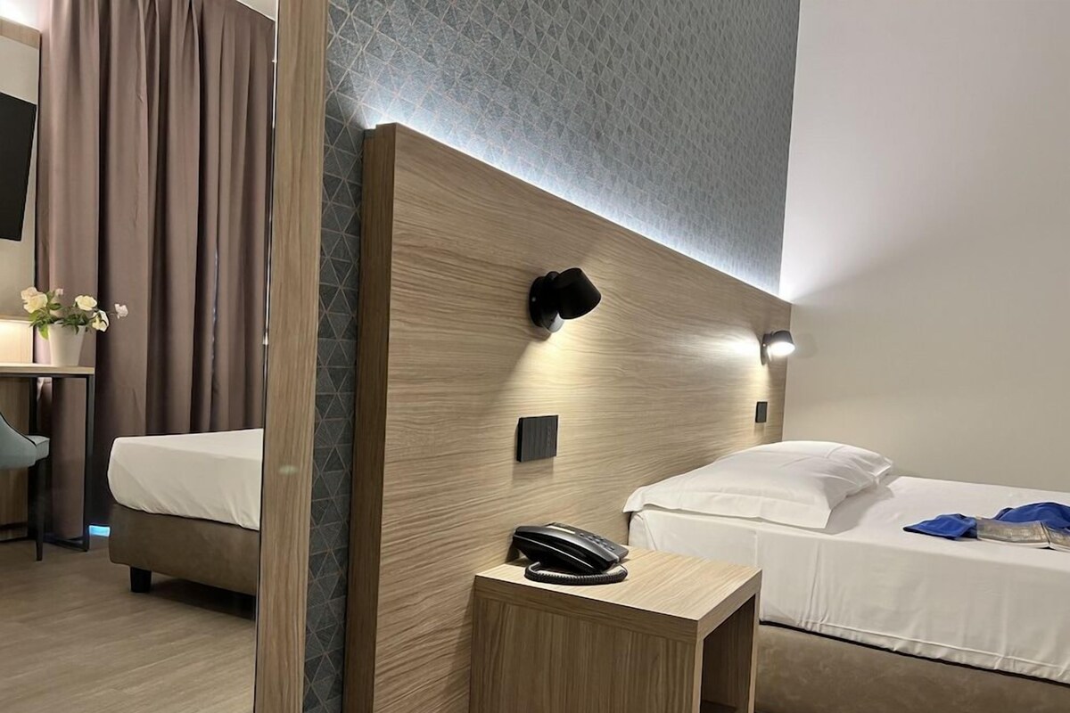 Habitación de hotel con cabecera de madera, mesita de noche con teléfono, cama con ropa blanca y una pequeña mesa con un jarrón de flores.