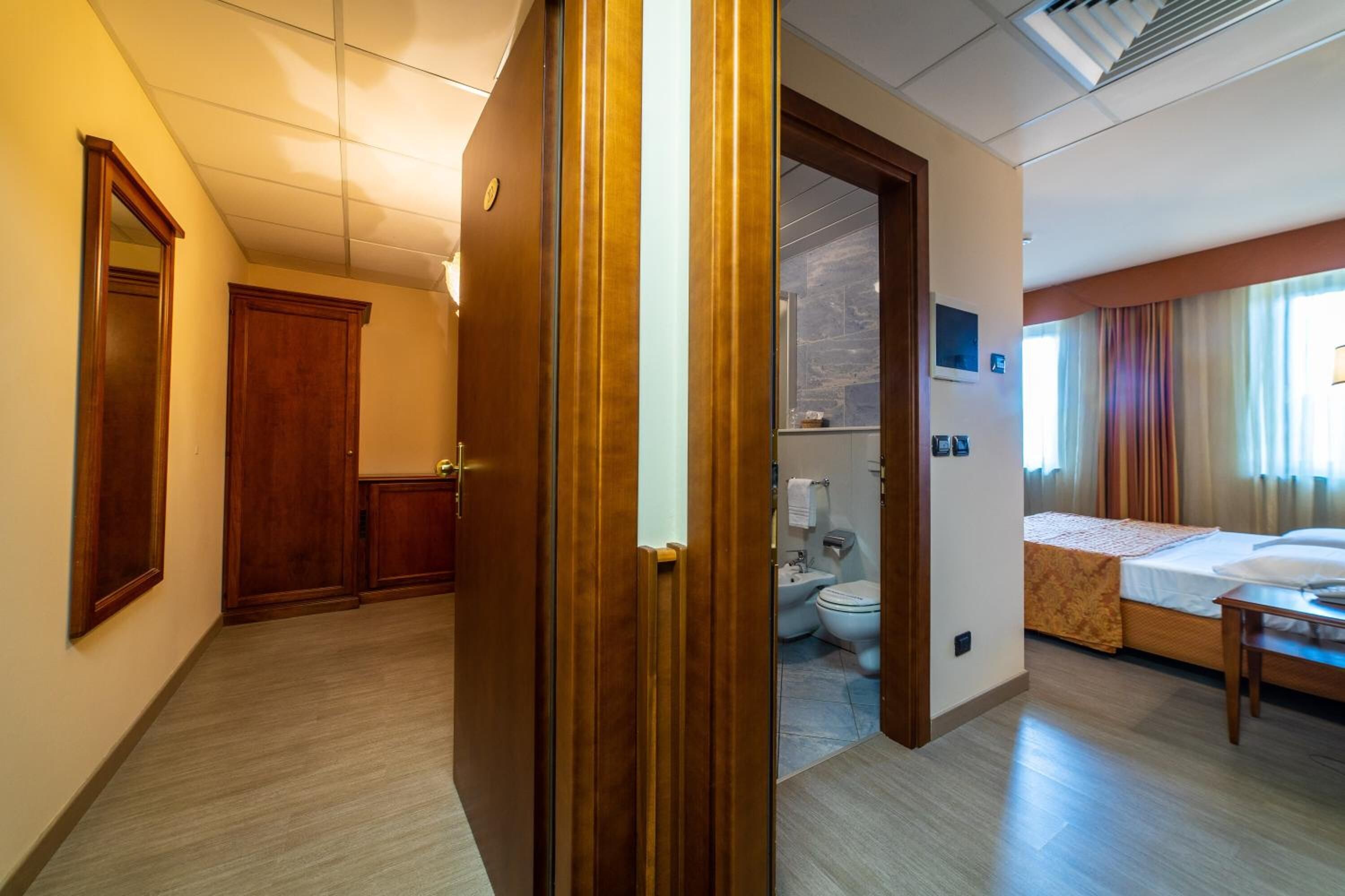 Una camera d'albergo con una porta in legno, un letto, un comodino, un bagno con il water e un bidet, e vista all'esterno da una finestra.