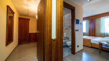 Una camera d'albergo con una porta in legno, un letto, un comodino, un bagno con il water e un bidet, e vista all'esterno da una finestra.